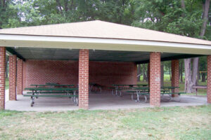 A brick picnic shelter over a concrete patio with six green picnic tables under it. Three sides are open, and the far end is a brick wall.