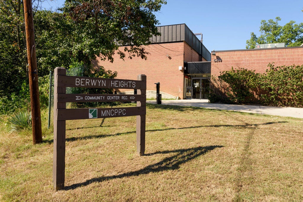 A brown wooden sign reading BERWYN HEIGHTS COMMUNITY CENTER in a grassy lawn with an arrow pointing to a large brick building.