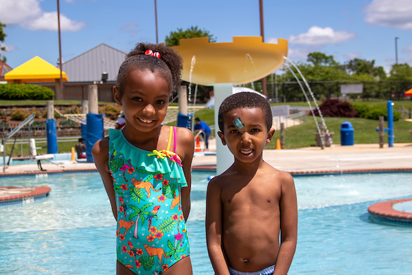 Two children in swimming suits smile while standing in front of a large pool with fountain and splash park elements, including a yellow umbrella fountain.