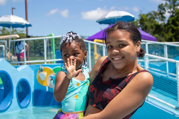A smiling young woman holds a small child while standing in front of a pool with colorful water park elements. Both are wearing colorful swimsuits.