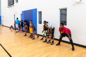 Young men stand in a line along one wall of a gymnasium, looking at the basketball court.