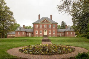 A brick path leads through a lawn to a large, Colonial-style brick mansion with a two-story central structure flanked by two single-story wings.