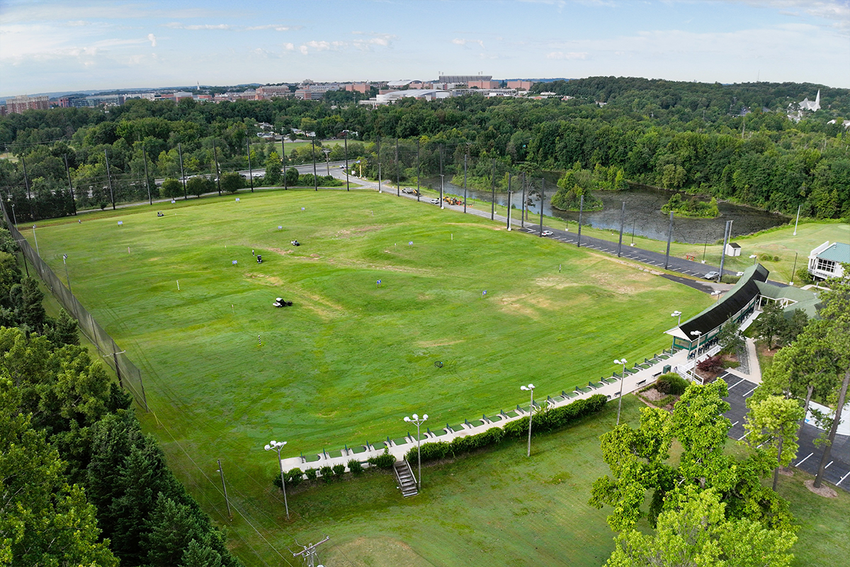 Aerial view of a driving range, featuring a large rectangular field of green grass with a line of several dozen teeing spots along the near end. The field is surrounded by a tall fence on three sides. Beyond the fence on the far side is a pond.