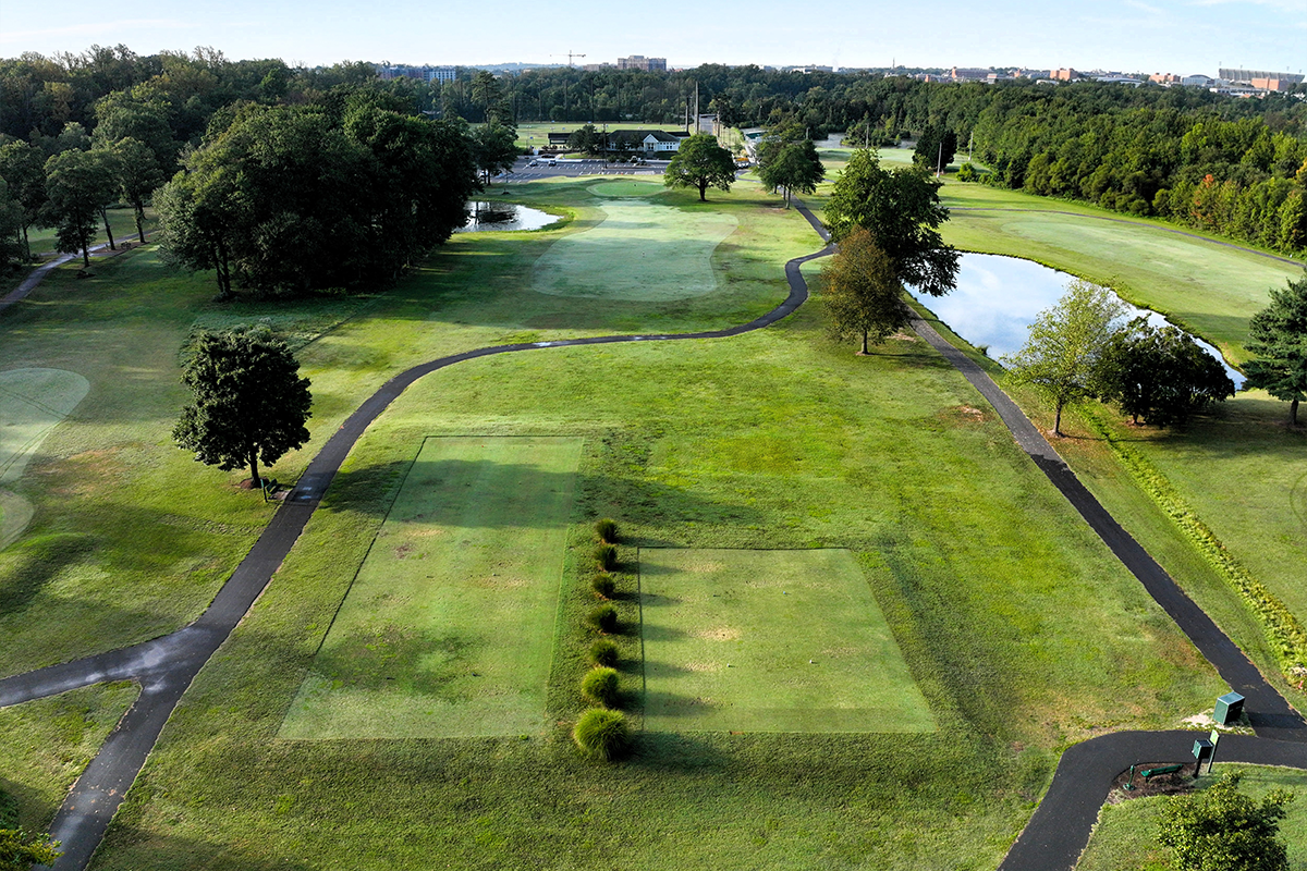 Aerial view of a hole on a golf course, featuring a rectangular patch of grass at the tee, a long fairway with two small ponds on either side, and a green circular hole in the distance, near a parking lot and a white building. A black, asphalt path winds around the entire course.