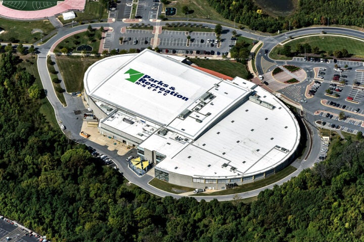 Aerial view of a large oval-shaped building with a white roof, emblazoned with a giant Parks and Recreation logo. There are several parking lots next to it on one side, and a wooded area on the other side.