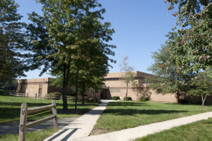Sidewalks leading through grass to a large brick building partially obscured by trees.