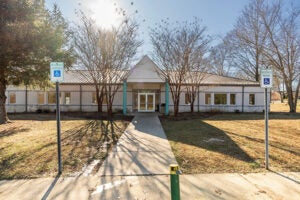Exterior view of the entrance of a long beige building. Over the double doors is a portico supported by two bright turquoise columns.