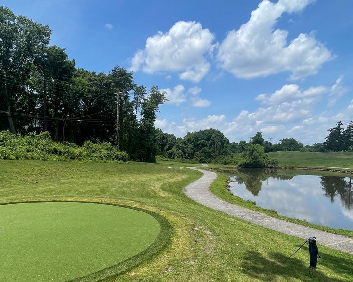A narrow paved path follows the edge of a body of water. On the other side of the path is a circular putting green.