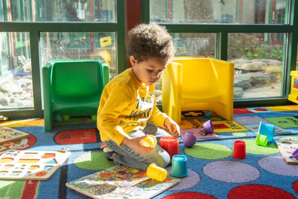 A small child plays with stacking cups on a colorful rug next to large windows. Near the child are wooden puzzled and plastic youth chairs.