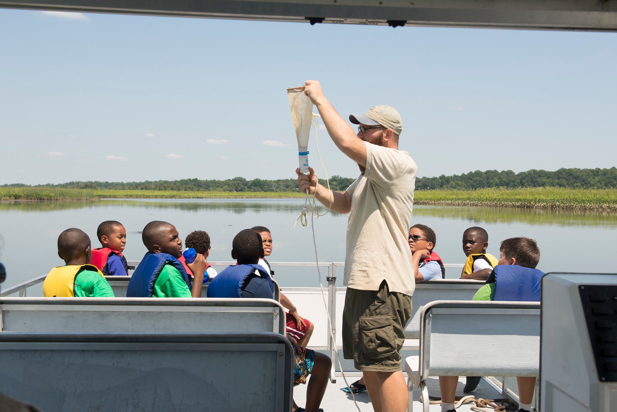 About ten kids in life vests sit on benches in the sun on a pontoon boat, all turning to look at a man in a staff t-shirt who is holding up a funnel attached to a thin rope.