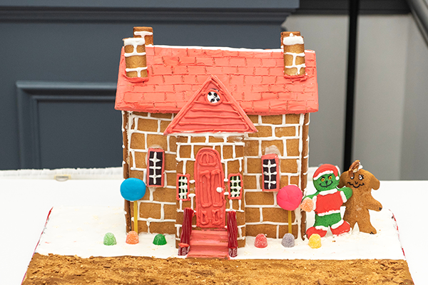 A quaint gingerbread house with frosting lined brickwork, frosting roofing, beautiful licorice hand rails, and wonderfully kept gumdrop shrubbery. The gingerbread Grinch and his gingerbread dog Max are standing to the side of the building. They are scheming something.