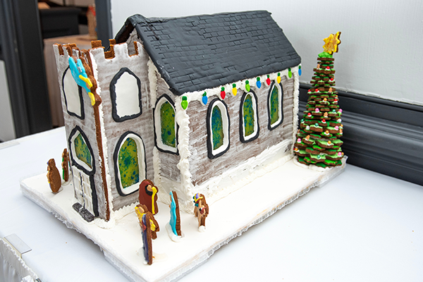 A white gingerbread church with white icing mortar and molding to hold it together. It has a large front steeple, as well as lively green and blue sugar stained glass windows throughout. There is a gingerbread nativity scene out front, and a large gingerbread tree at the back corner of the building.
