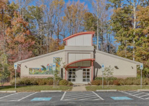 Exterior view of a beige building with a peaked roof and a peach portico over the front doors. There is a colorful mural on one wall.