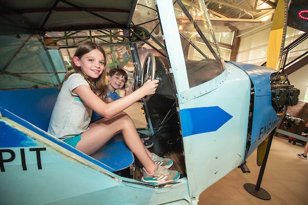 Two kids sit in the pilot seats of a vintage, blue aircraft, holding onto the silver metal "steering wheels" in front of them.