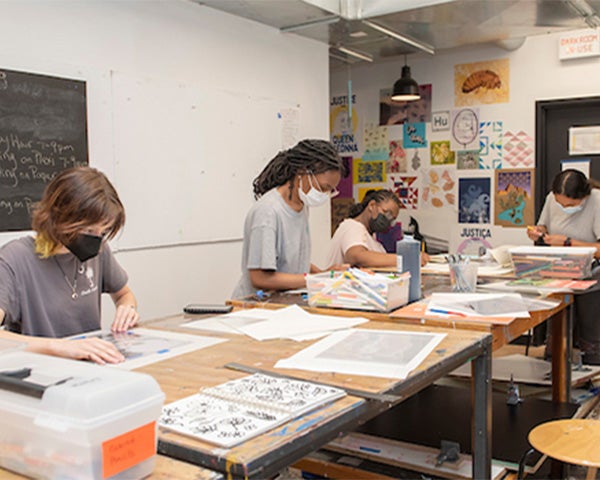 In a classroom, four young women wearing masks work at large wooden tables on art projects. There are several dozen artworks hanging on the far wall.