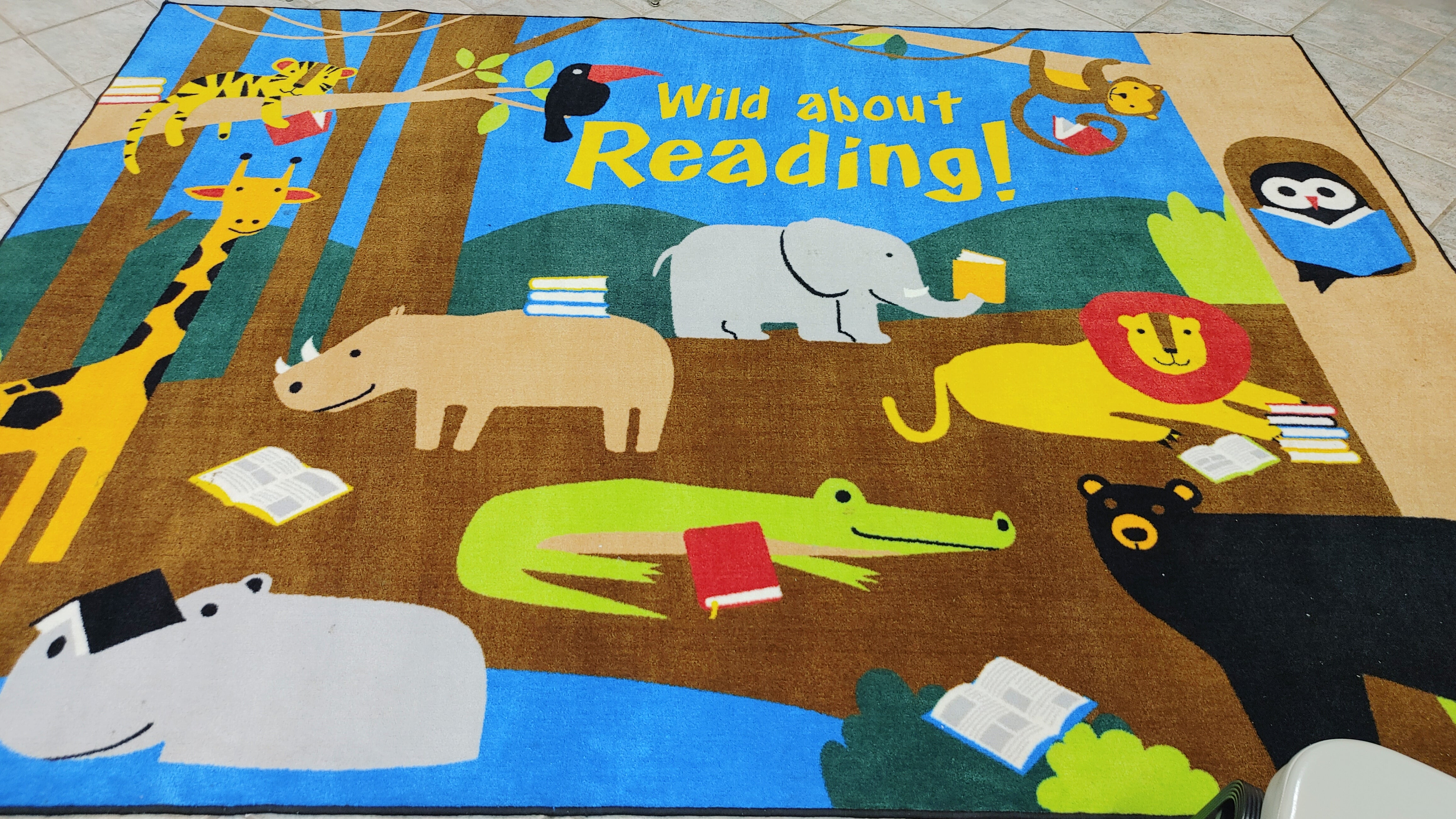 A colorful rug on a beige, tiled floor, with cartoonish images of wild animals in a nature setting with stacks of books, and the words WILD ABOUT READING at the top.