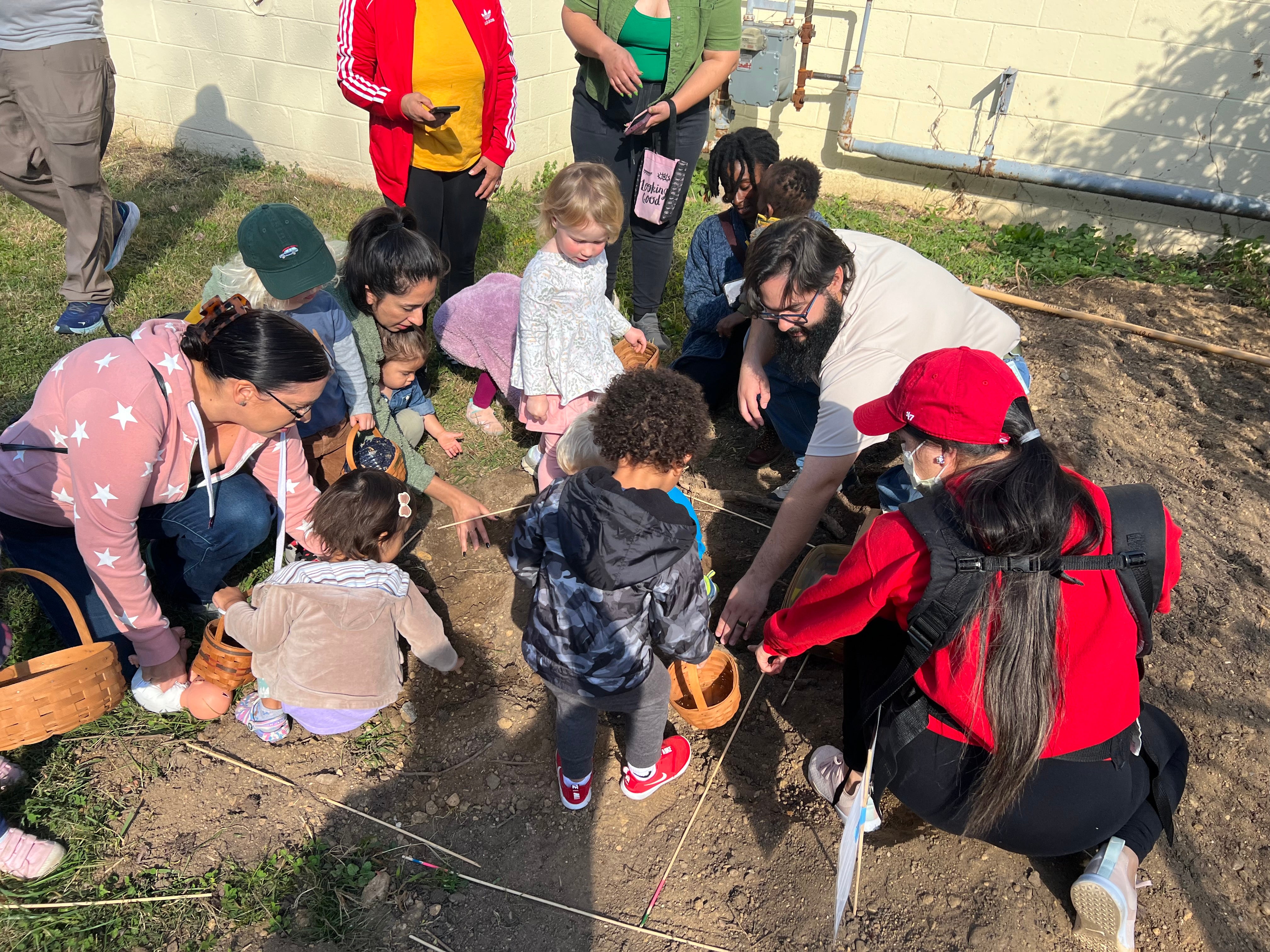 Outside, about five toddlers and their parents sit and stand on a large patch of dirt in a lawn next to a beige building. The toddlers hold small baskets.
