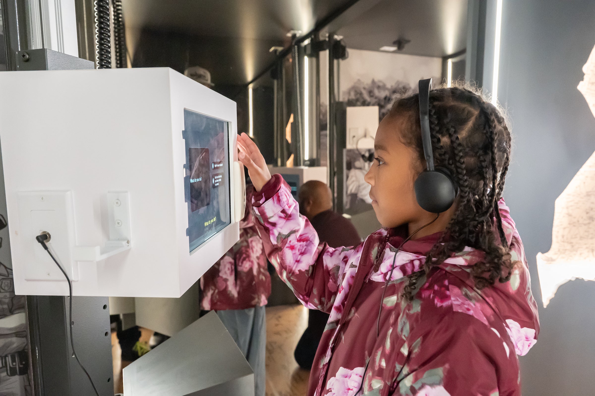 A girl in a floral coat interacts with a video monitor museum exhibit, while wearing headphones connected to the exhibit. Behind her other visitors explore the museum.