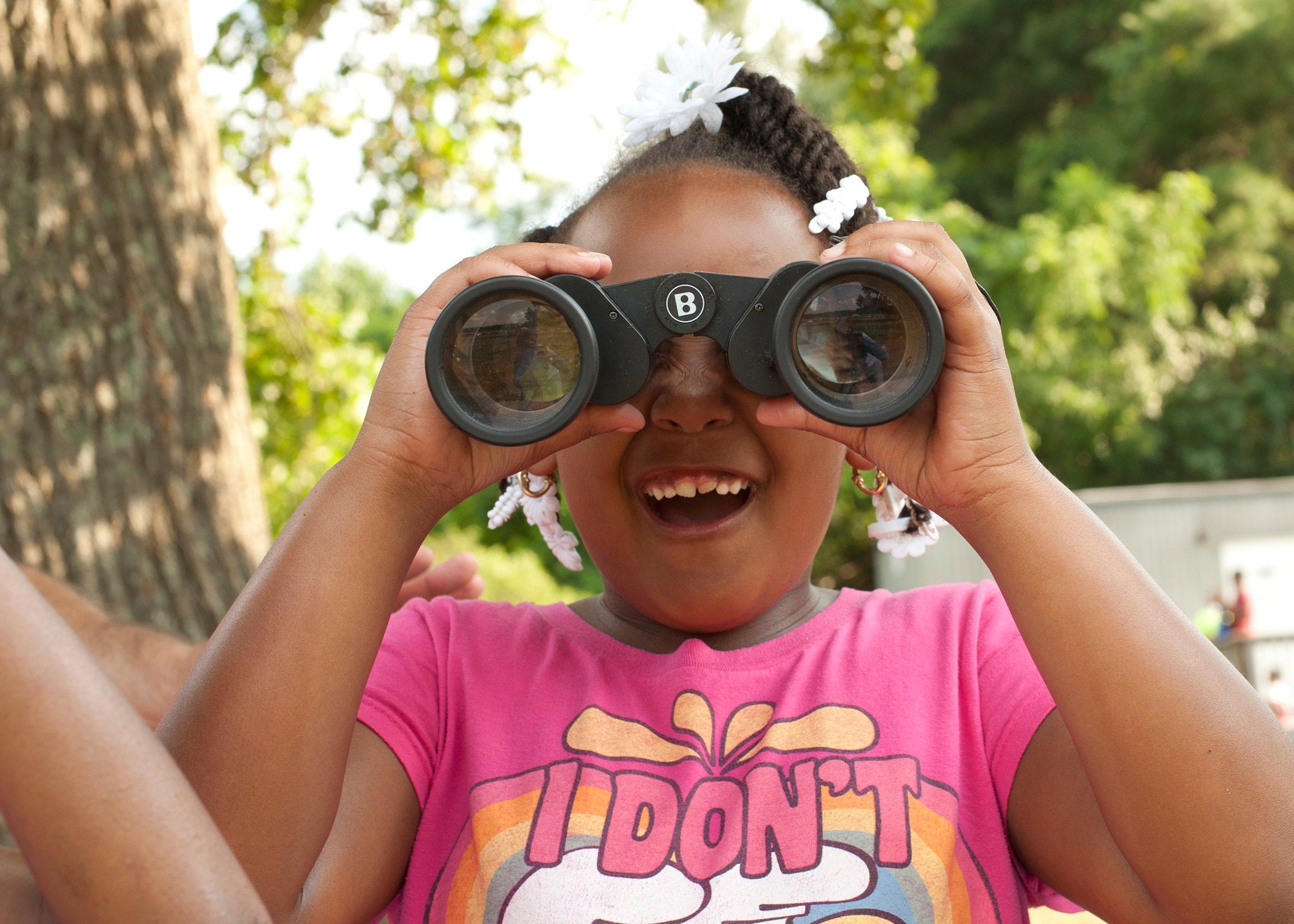 A child in a pink t-shirt uses black binoculars to look directly at the photographer of the image. Her mouth is open in an expression of delight. Behind her is a blurry tree.