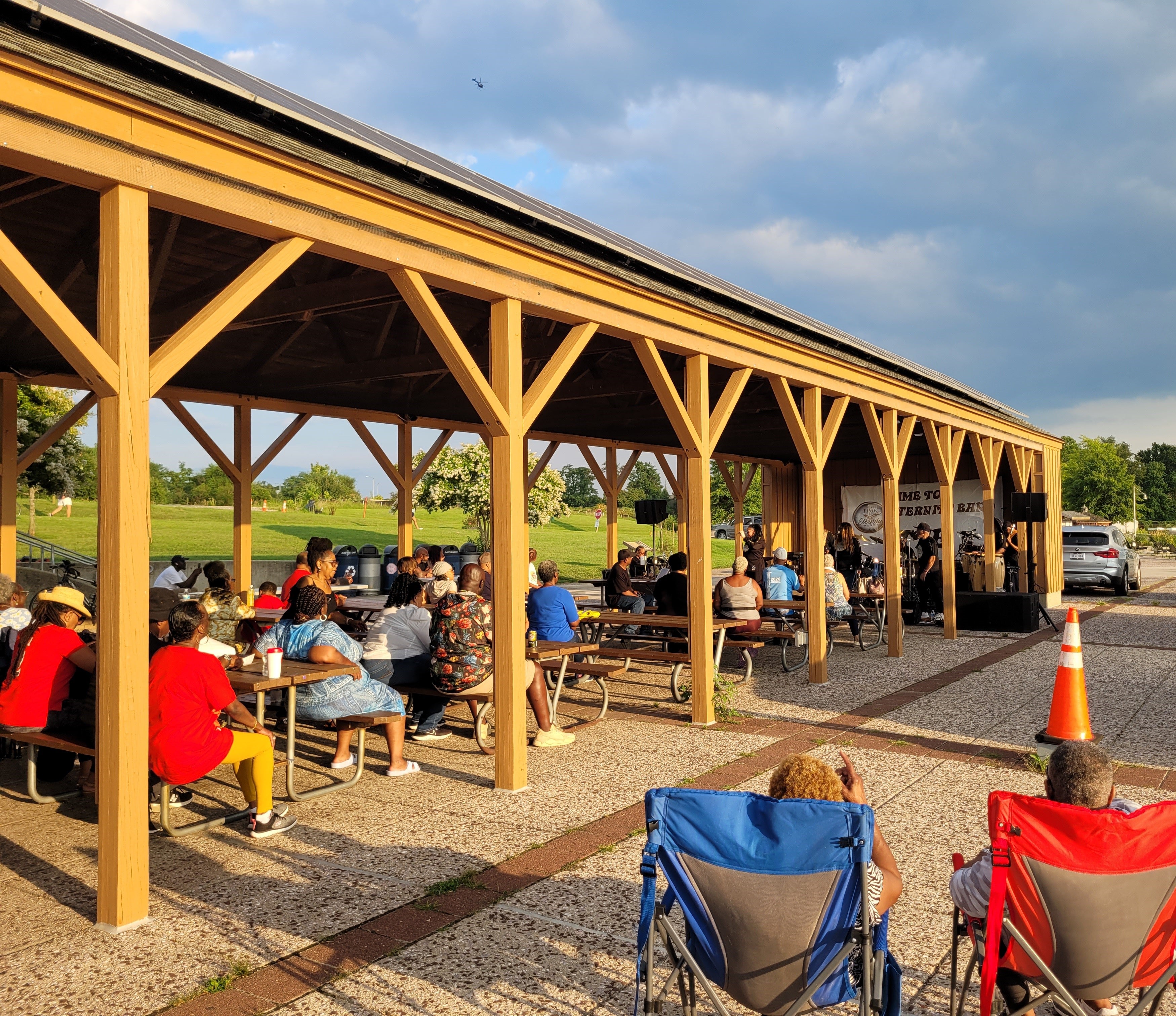 At sunset, spectators sit at picnic tables under a long, wooden, pavilion with a live band performing at the far end.