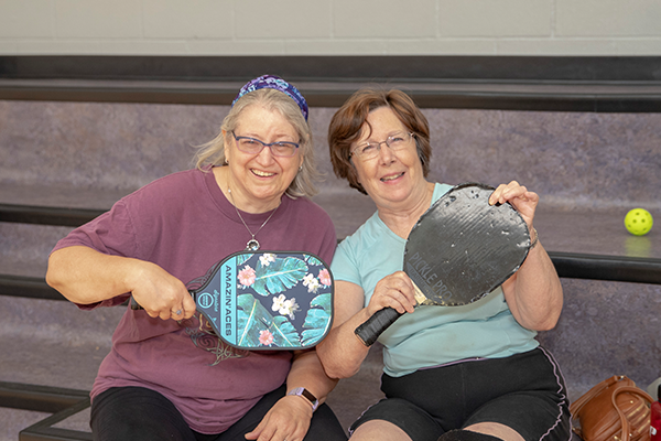 Two women sitting, posing while holding up pickleball paddles