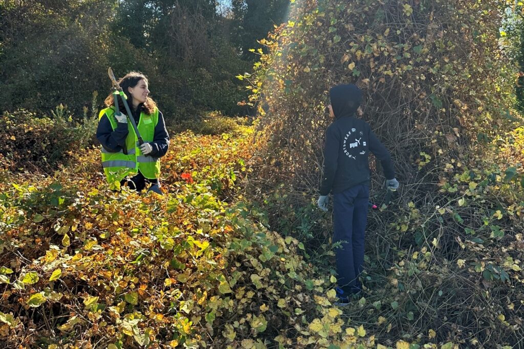 A picture of a woman and a young child in an area covered with vines. The woman is wearing gloves and a safety vest and is holding a pair of loopers.