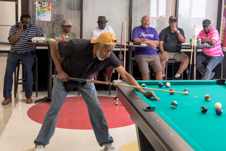 A man in a yellow hat lines up a shot on a billiard table. Behind him half a dozen men in tall stools look on. The linoleum floor has a decorative polka-dot pattern in primary colors.
