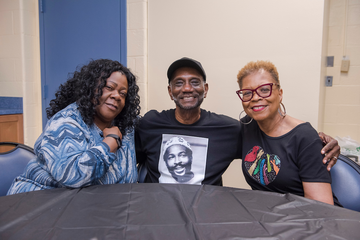 Three smiling friends sit with their arms around each other's shoulders, at a table with a black tablecloth. The man in the center is wearing a Marvin Gaye t-shirt.
