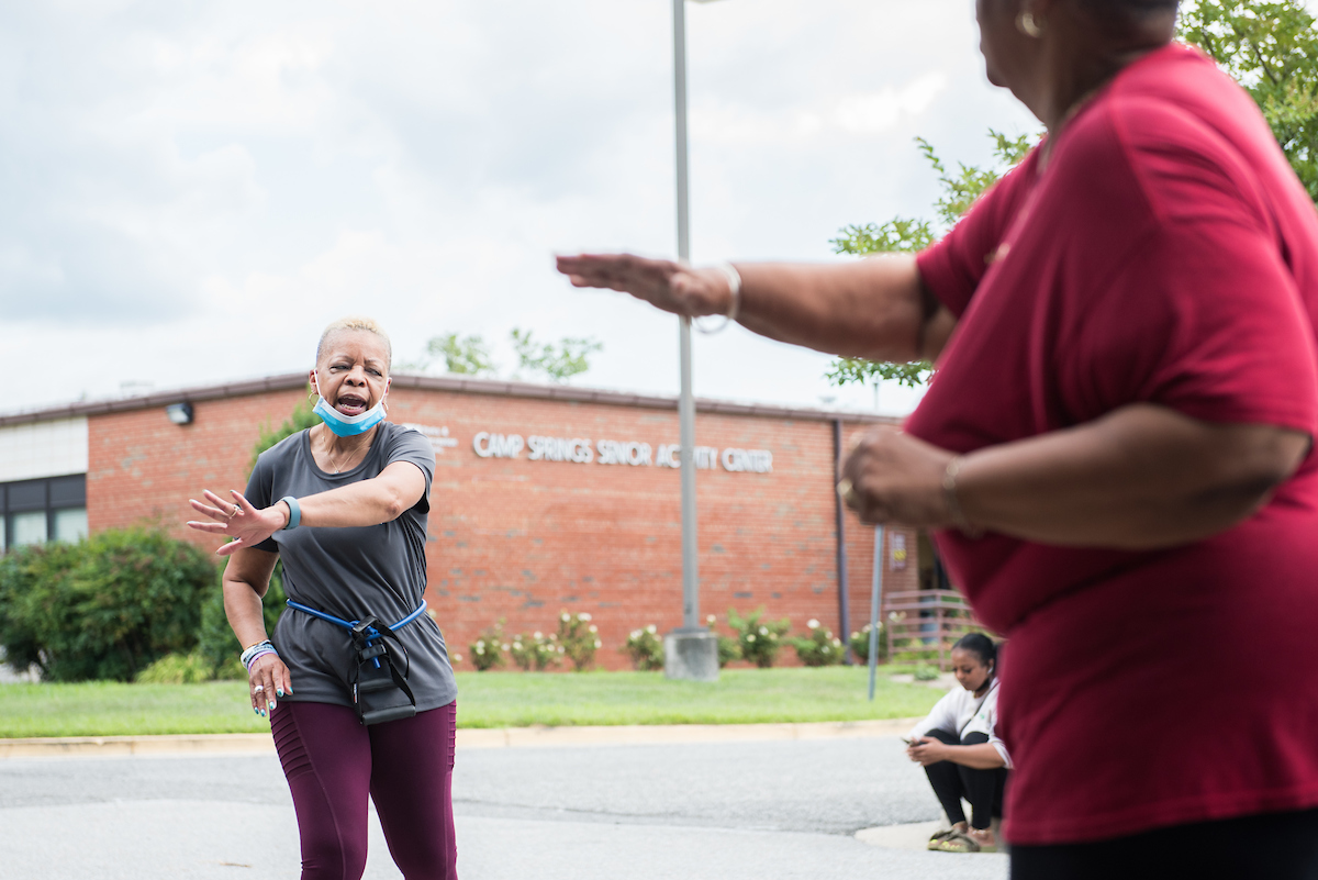 A fitness instructor demonstrates an arm stretch in front of a class of seniors in a parking lot. Behind them is a single-story brick building with the words CAMP SPRINGS SENIOR ACTIVITY CENTER.