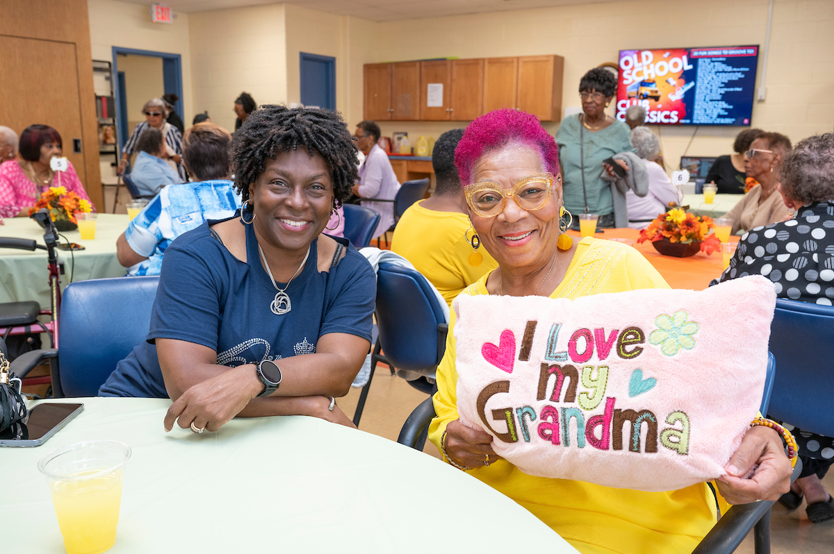 Two smiling senior women sit together at a banquet table in a large room. One holds a colorful pillow that reads "I love my grandma." Behind them in the room are dozens more seniors seated at banquet tables.