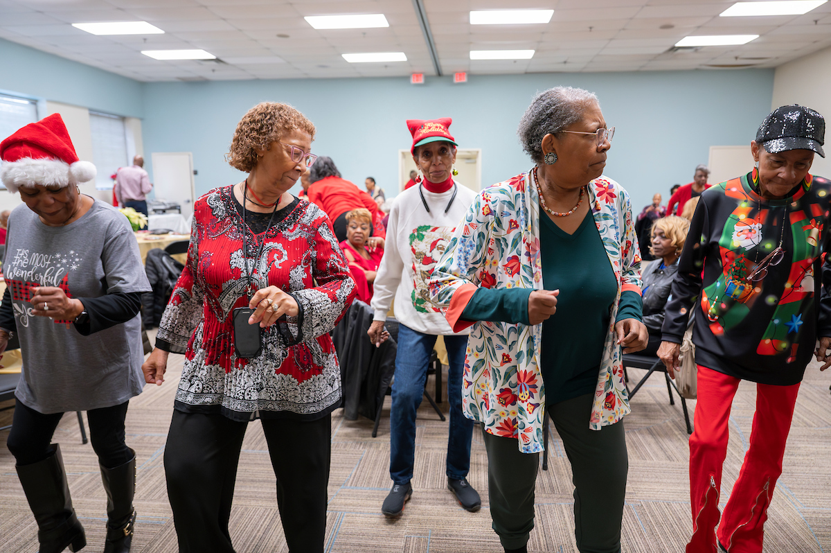 Five senior women wearing holiday-themed outfits dance in a large event room with blue walls. Behind them are tables where more seniors are seated, all in festive holiday clothing.