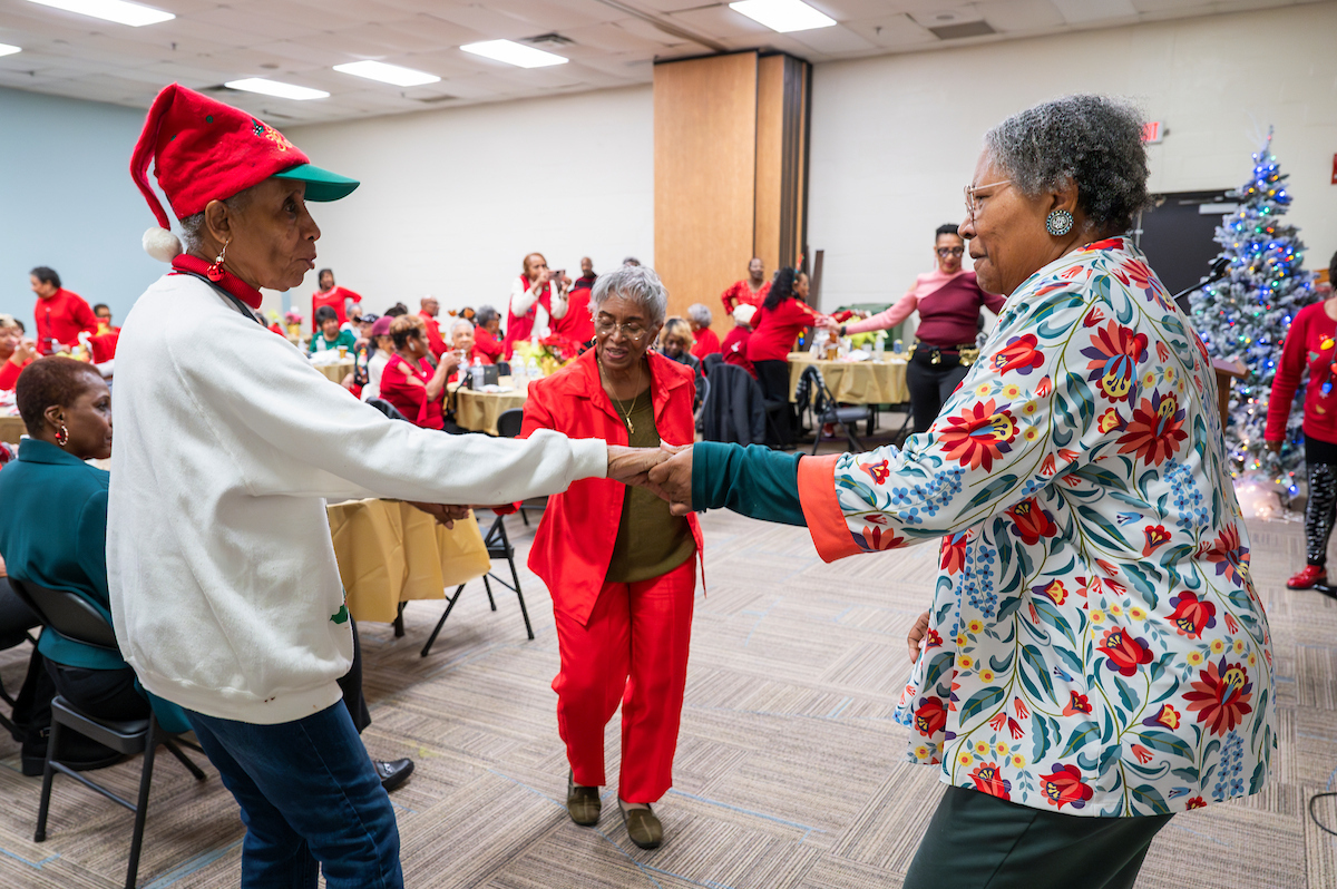 Three senior women dance while holding hands in a circle. Behind them are banquet tables with more seniors, and in the far corner is a decorated holiday tree. Everyone in the room has a festive holiday outfit on.