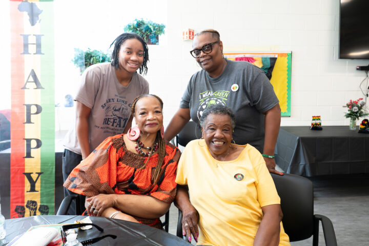 Two staff members stand behind two senior women seated at a banquet table. Behind them the room is decorated with colorful red, yellow, and green Juneteenth holiday banners.