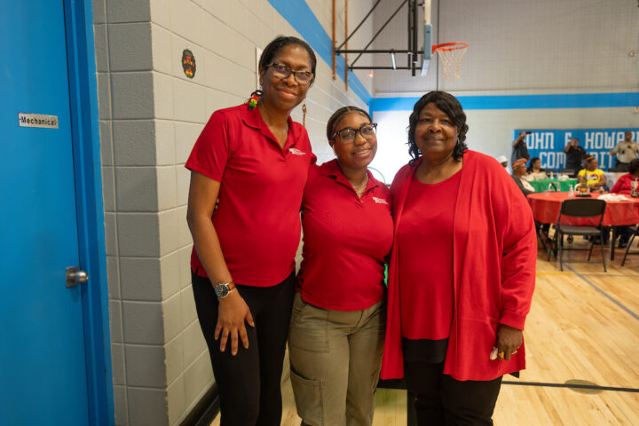 Three women in red shirts stand smiling in a gymnasium. Two are wearing glasses and Parks and Recreation staff shirts. Behind them, the gym is set up with tables and chairs for a party.