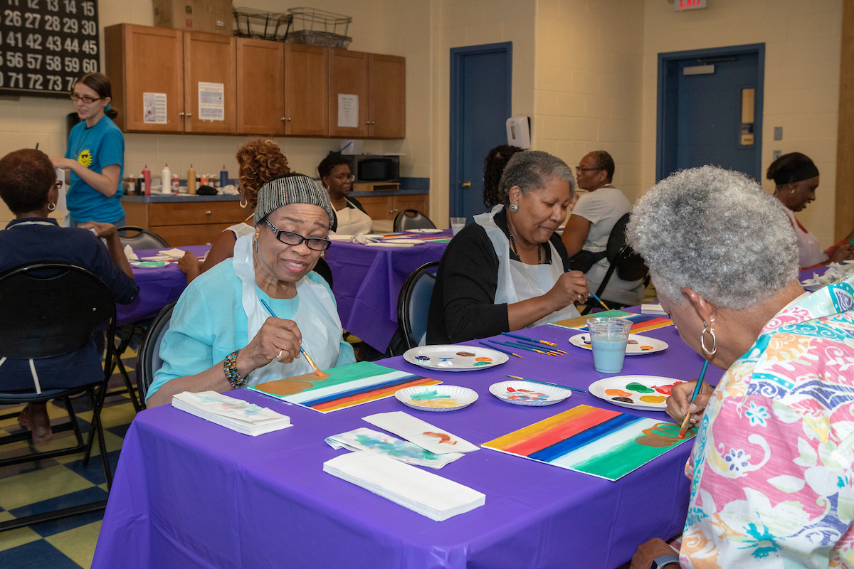 Seniors sit at long tables painting colorful stripes onto canvases. In the back of the room is a young staffer giving instructions. The seniors wear plastic smocks and the tables have protective purple coverings.