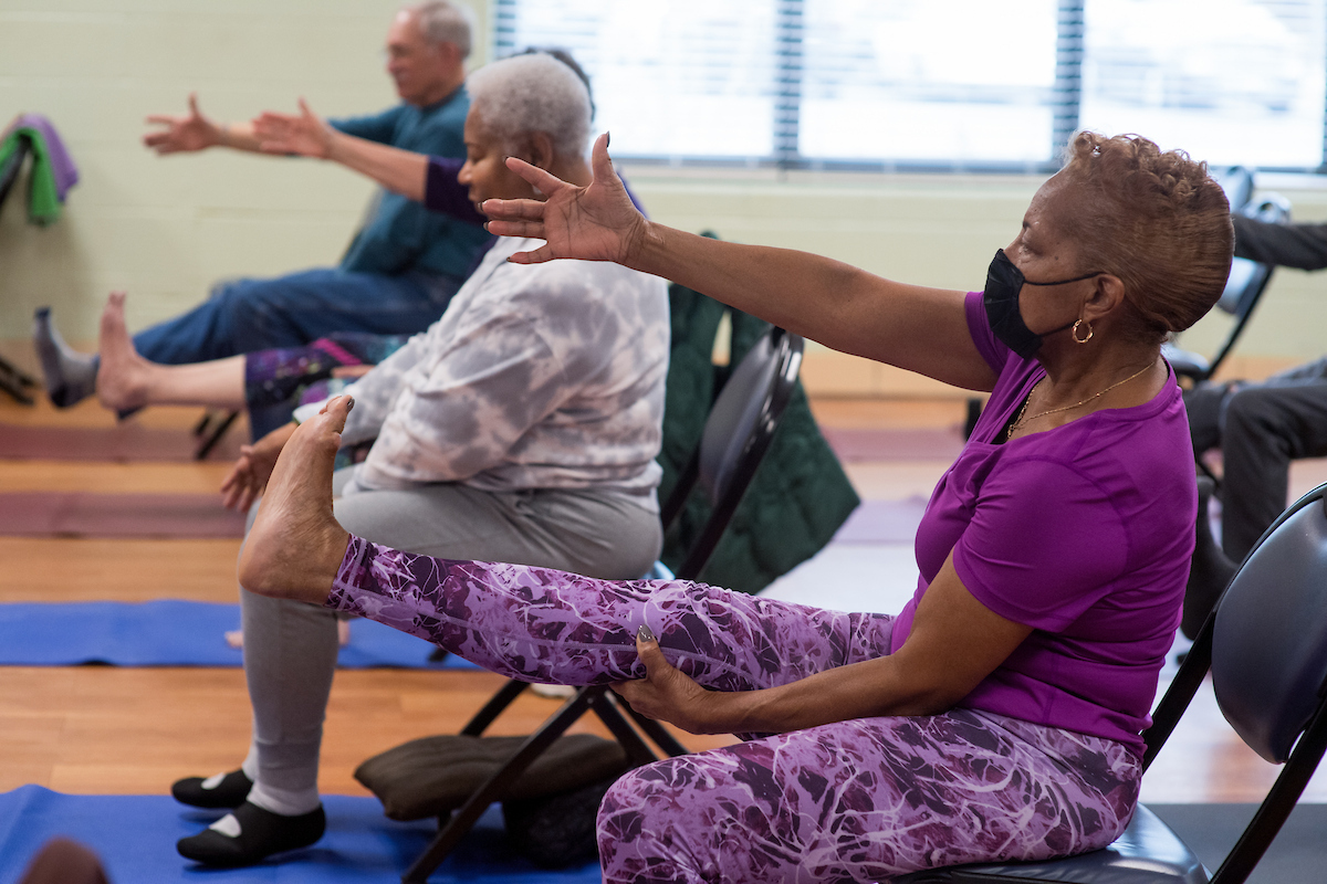 Senior men and women in comfortable workout clothing sit in rows of folding chairs atop yoga mats in a room with a wooden floor. They are all extending their right arms and legs forward in a yoga pose.