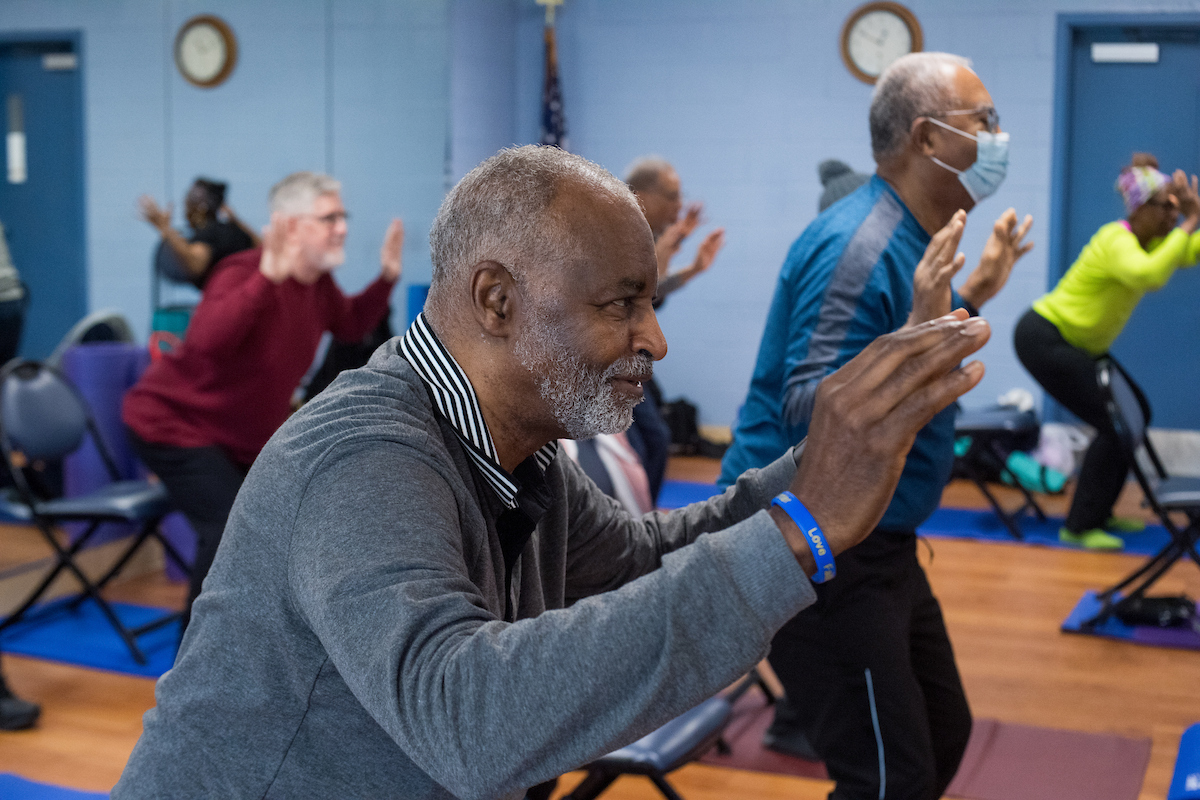 Senior men and women in comfortable workout clothing stand among rows of folding chairs atop yoga mats in a room with a wooden floor. They all have their arms slightly raised while crouching in a yoga pose.