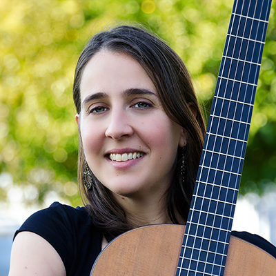 A woman smiling holding an acoustic guitar against a blurry tree line background