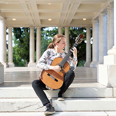 A person sitting on marble stairs surrounded by pillars playing their acoustic guitar