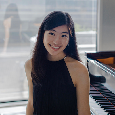 A woman sitting on the bench of a grand piano smiling at the camera against a blurry window leading outdoors