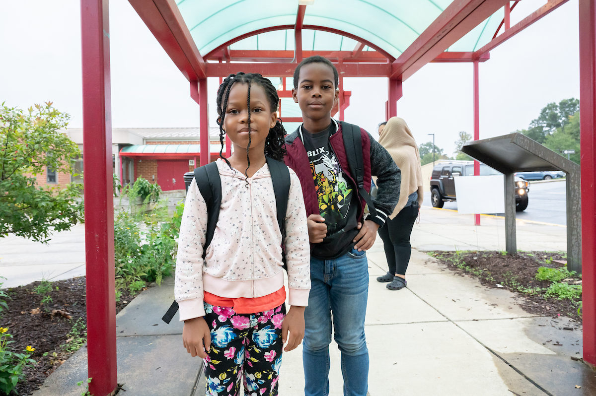 Two children wearing backpacks stand outside on a sidewalk under a red and green awning. There is a low brick building behind them.