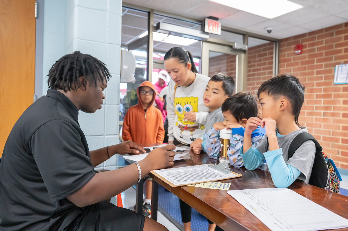 Near the glass doors inside a brick and cinderblock lobby, a woman stands at a check-in table with her three sons beside her. A man in a staff polo shirt sits behind the table with a clipboard.