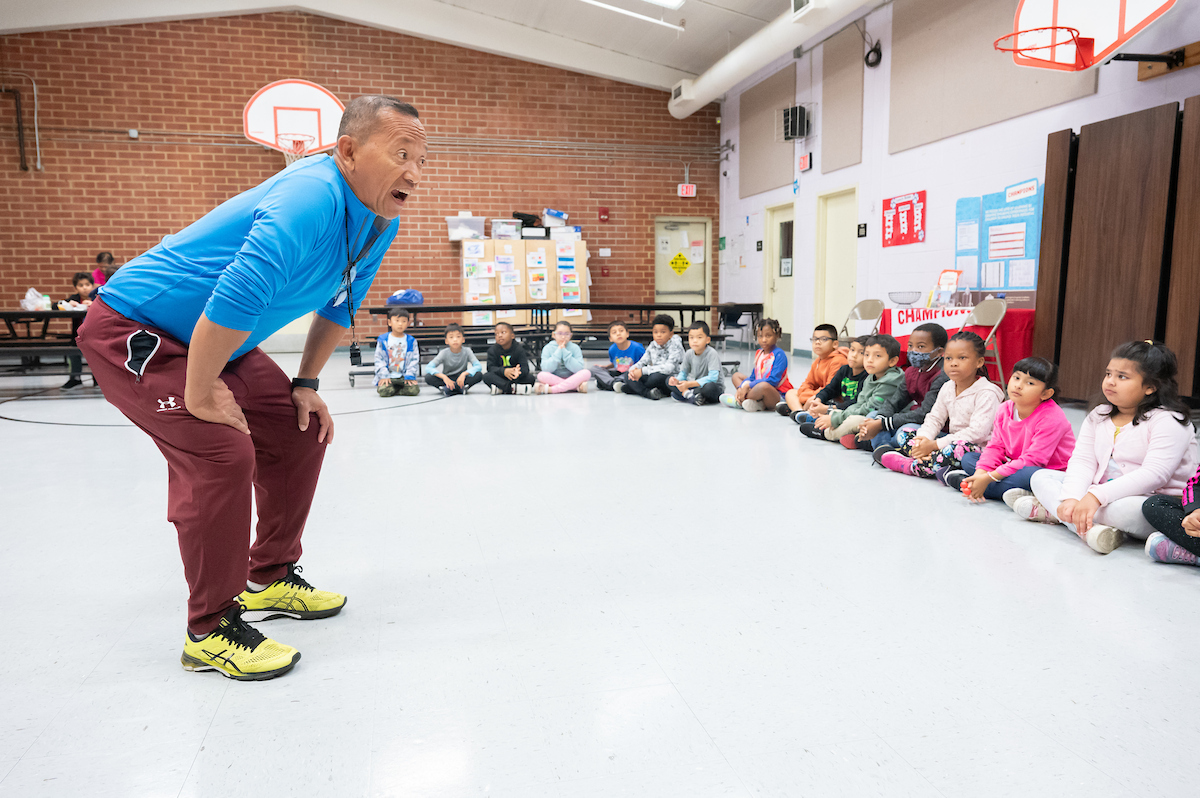 In a large room with basketball goals and lunch tables, an adult leans forward to address a group of children seated in a semi-circle in front of him. The man wears athletic clothes and has a whistle around his neck.