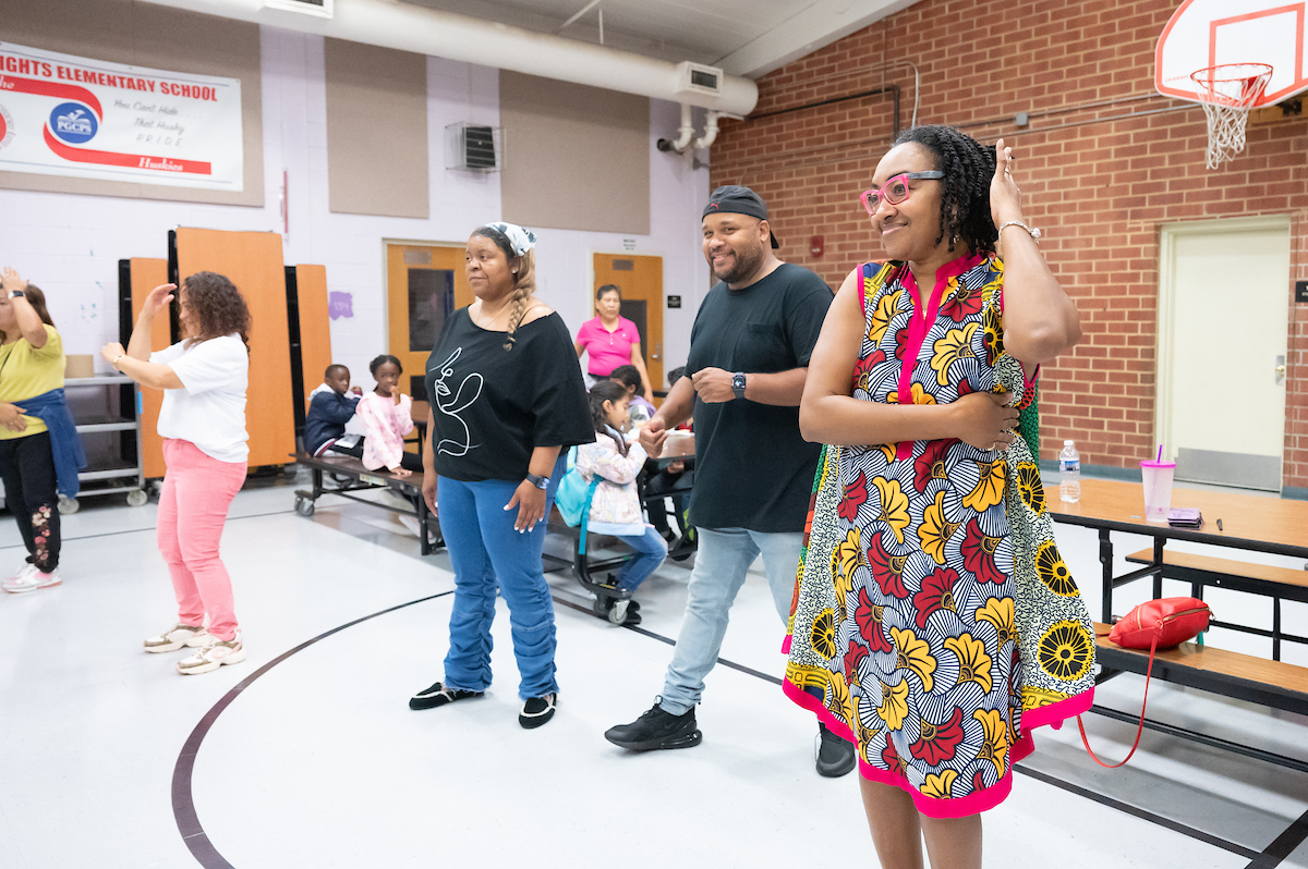 Five smiling adults stand in a combination gymnasium/lunch room doing the arm motions of a dance. Behind them are several long tables with children seated at them. There is a basketball goal on one wall, and the white linoleum floor has basketball court markings.