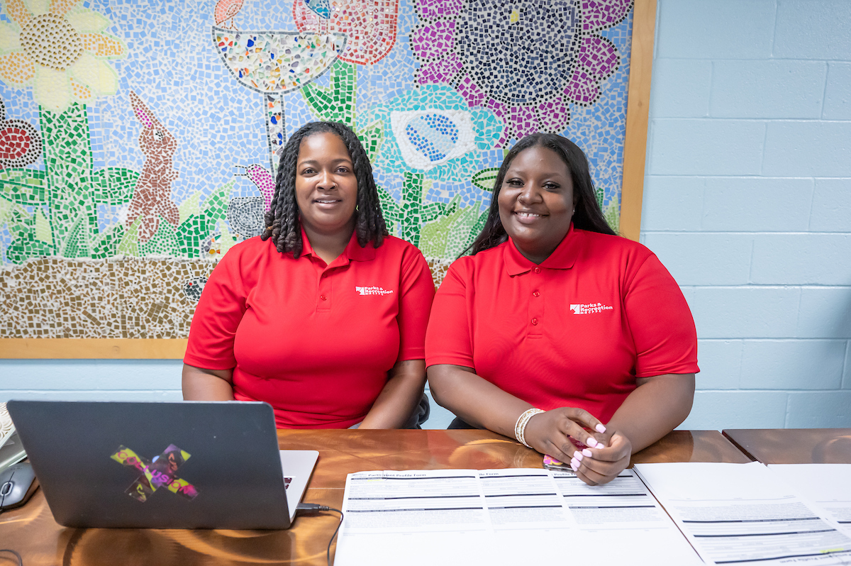 Two smiling women in red Parks and Recreation staff shirts sit at a check-in table. On the table are some papers and a laptop. Behind them is a light blue cinderblock wall with a large, colorful mosaic of flowers hanging on it.