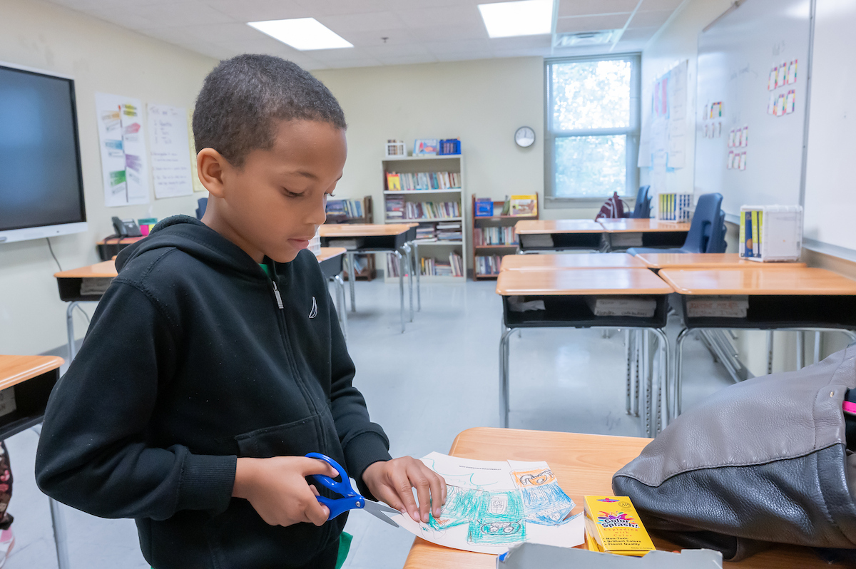 In a classroom with clusters a desks, a child uses scissors to cut a paper project that has been colored with crayons.