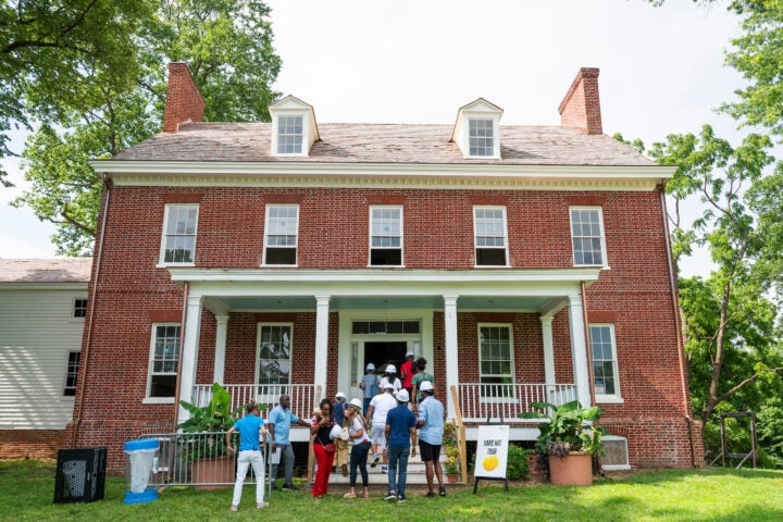 About 20 people in hard hats climb the porch steps to enter a two-story, Colonial brick building. A sign in the grass reads HARD HAT TOUR.