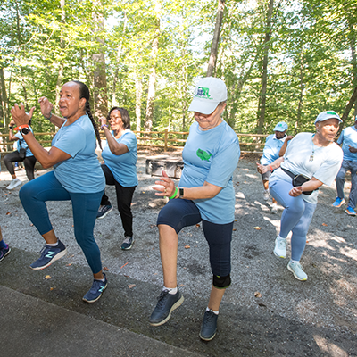 A group of people out on a trail doing exercises. They are all wearing light blue shirts, and are in a synchronized position of raising their right left in a stepping motion. There is a gravel path, benches, and many trees in the background.