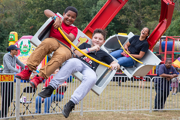 Three children sitting in a car on a carnival ride. They are spinning around in a circle. There are other people in the background and lots of other colorful carnival rides in the background