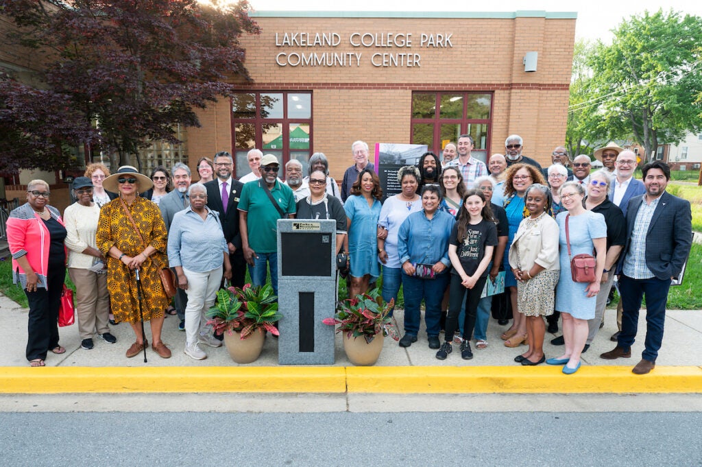 About 35 smiling people in front of a brick building with the words LAKELAND COLLEGE PARK COMMUNITY CENTER on it. The group stands behind a podium.